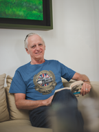 Man sitting on a couch wearing a blue t-shirt with a logo, in a room with a green screen in the background.