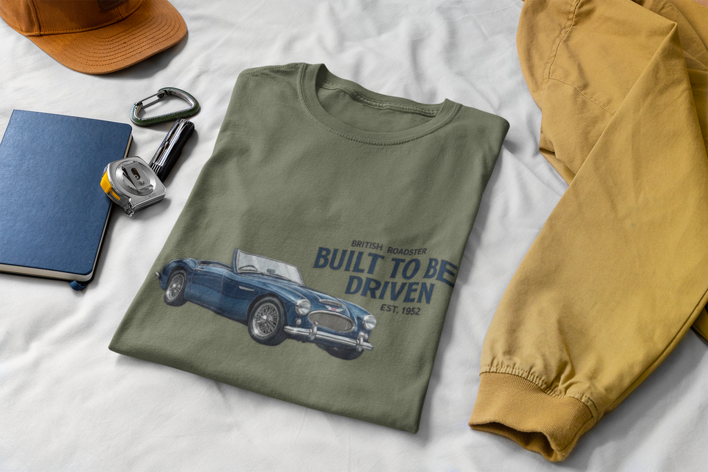 Folded military green t-shirt with Healey car graphic and text on a white surface with accessories.