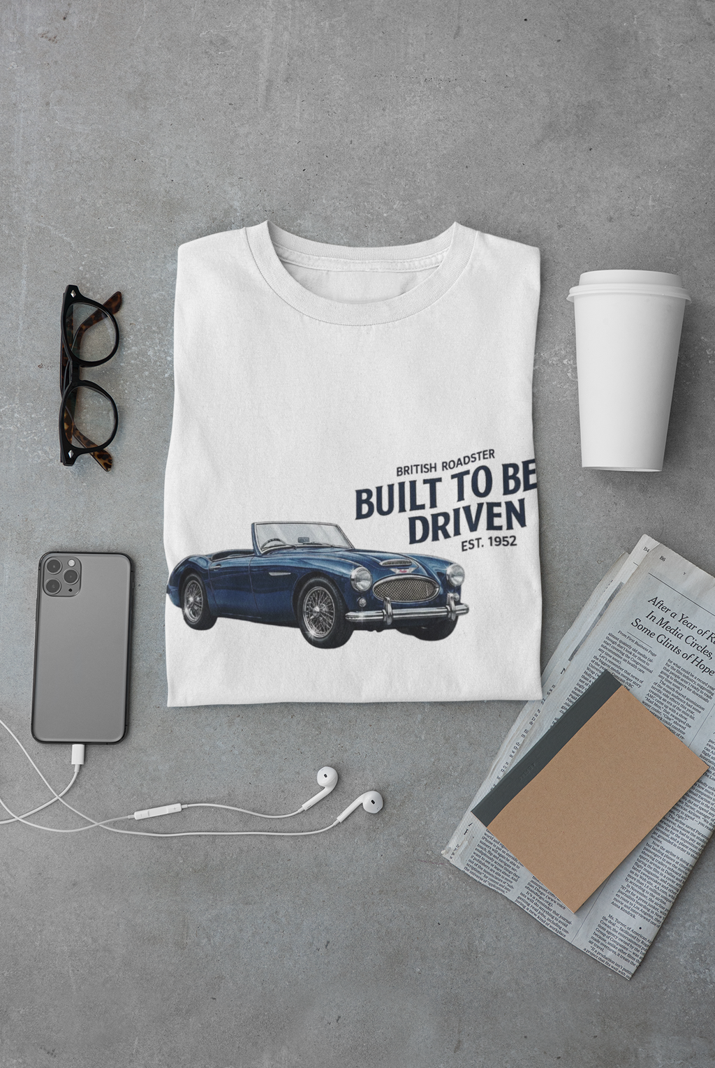 White t-shirt with classic Healey car graphic and text on a gray surface with accessories.