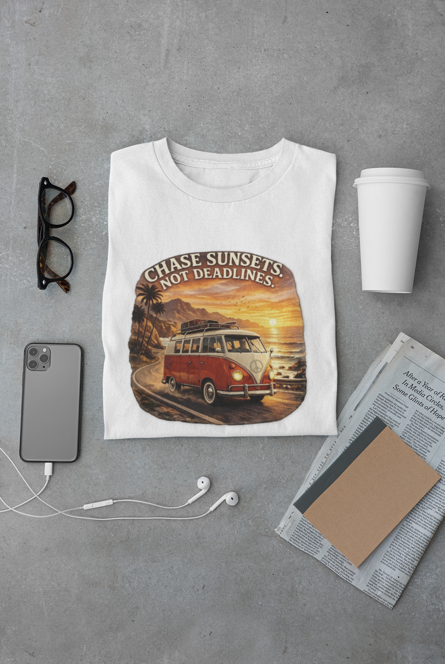 White t-shirt with a graphic design of a classic van and sunset text, placed on a gray surface with a phone, coffee cup, and glasses.