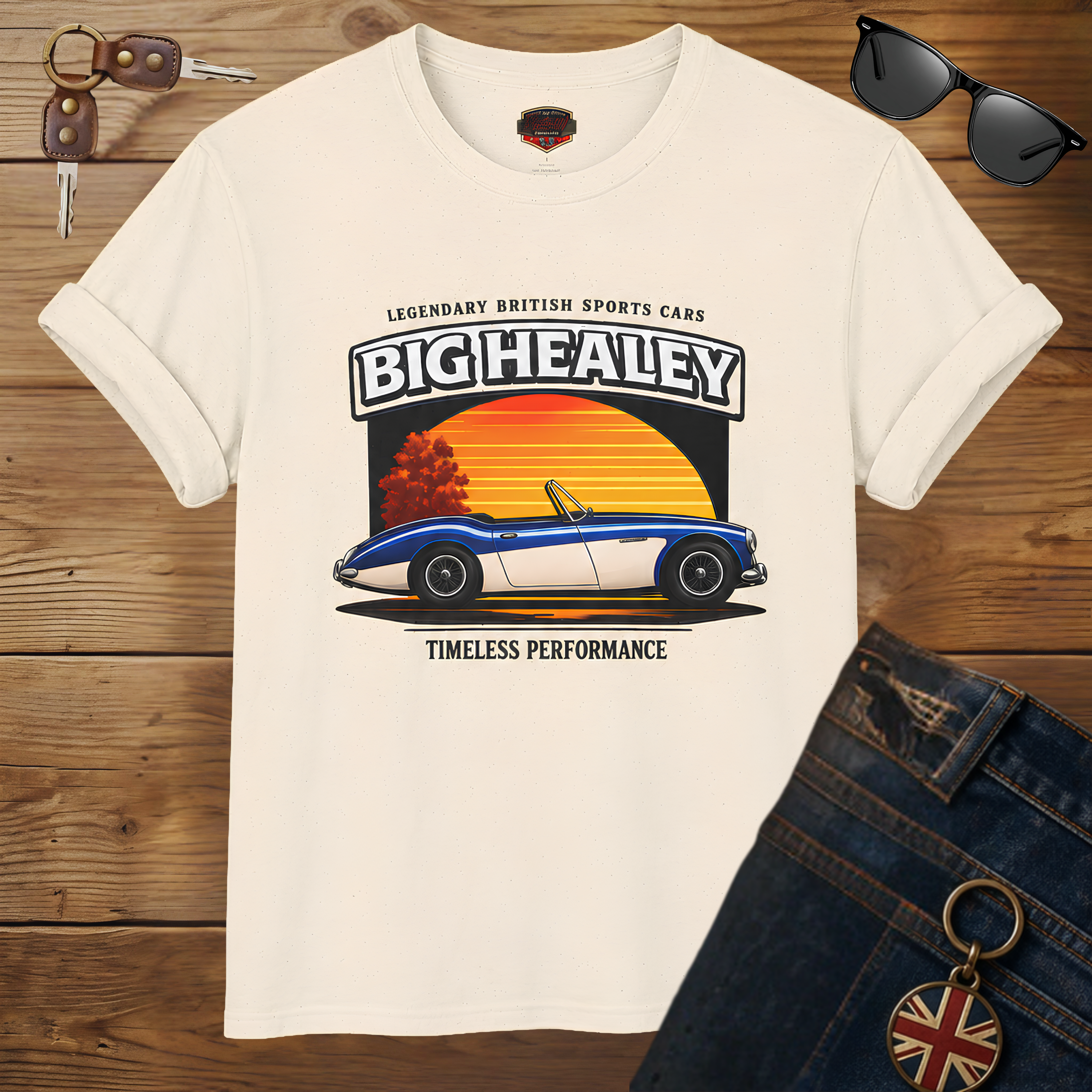 Big Healey graphic t-shirt featuring a blue and white Austin Healey roadster with retro sunset artwork and vintage British sports car typography on a wooden background.