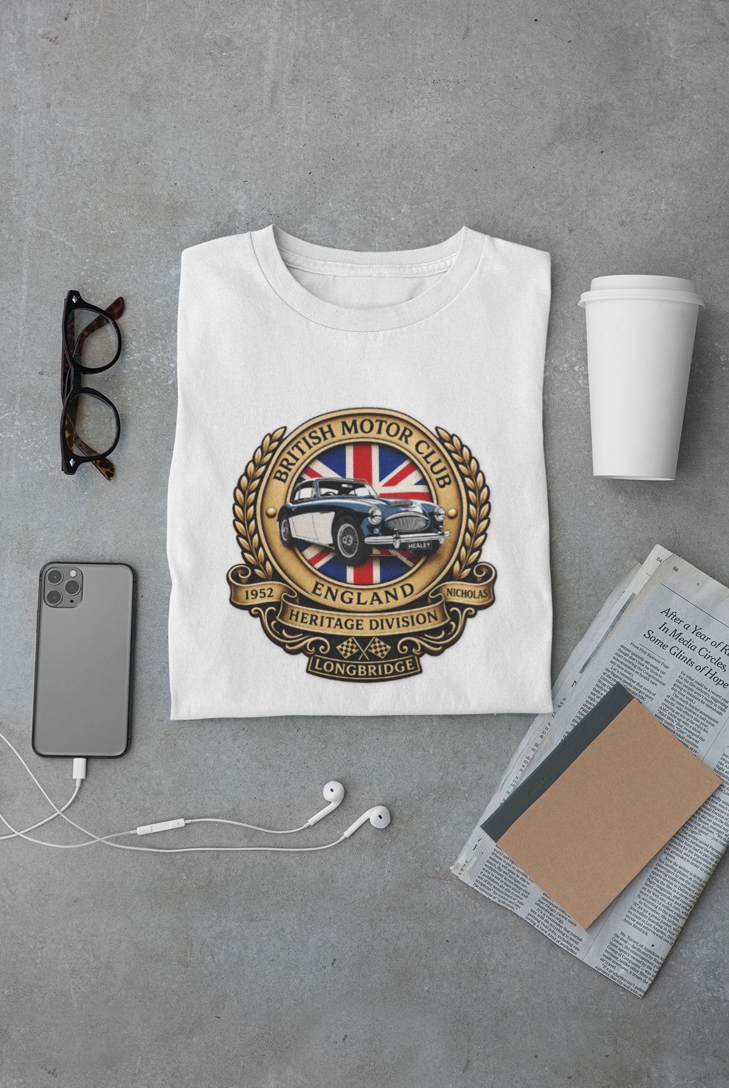 White t-shirt with a graphic design on a gray surface with a phone, cup, and glasses.