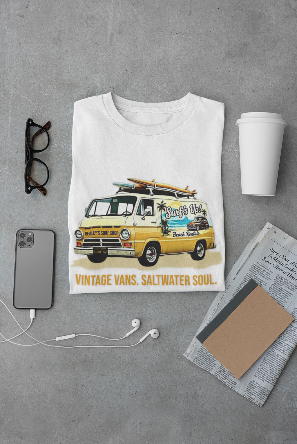 Folded white t-shirt with a classic van graphic and text on a gray surface with accessories.