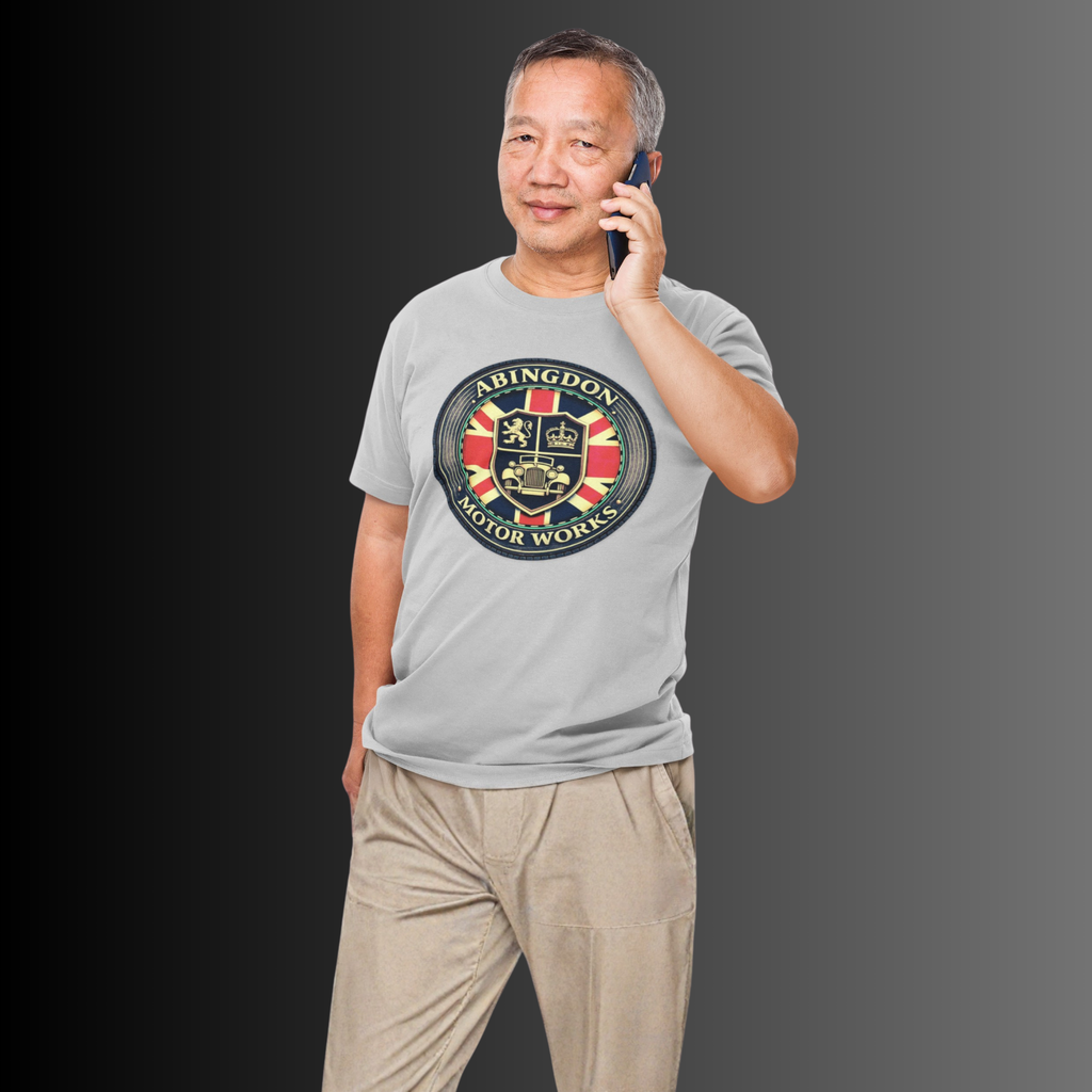 A man wearing a light gray t-shirt with a circular 'Abingdon Motor Works' crest graphic on the chest, paired with beige trousers, holding a phone to his ear against a dark gray gradient background.