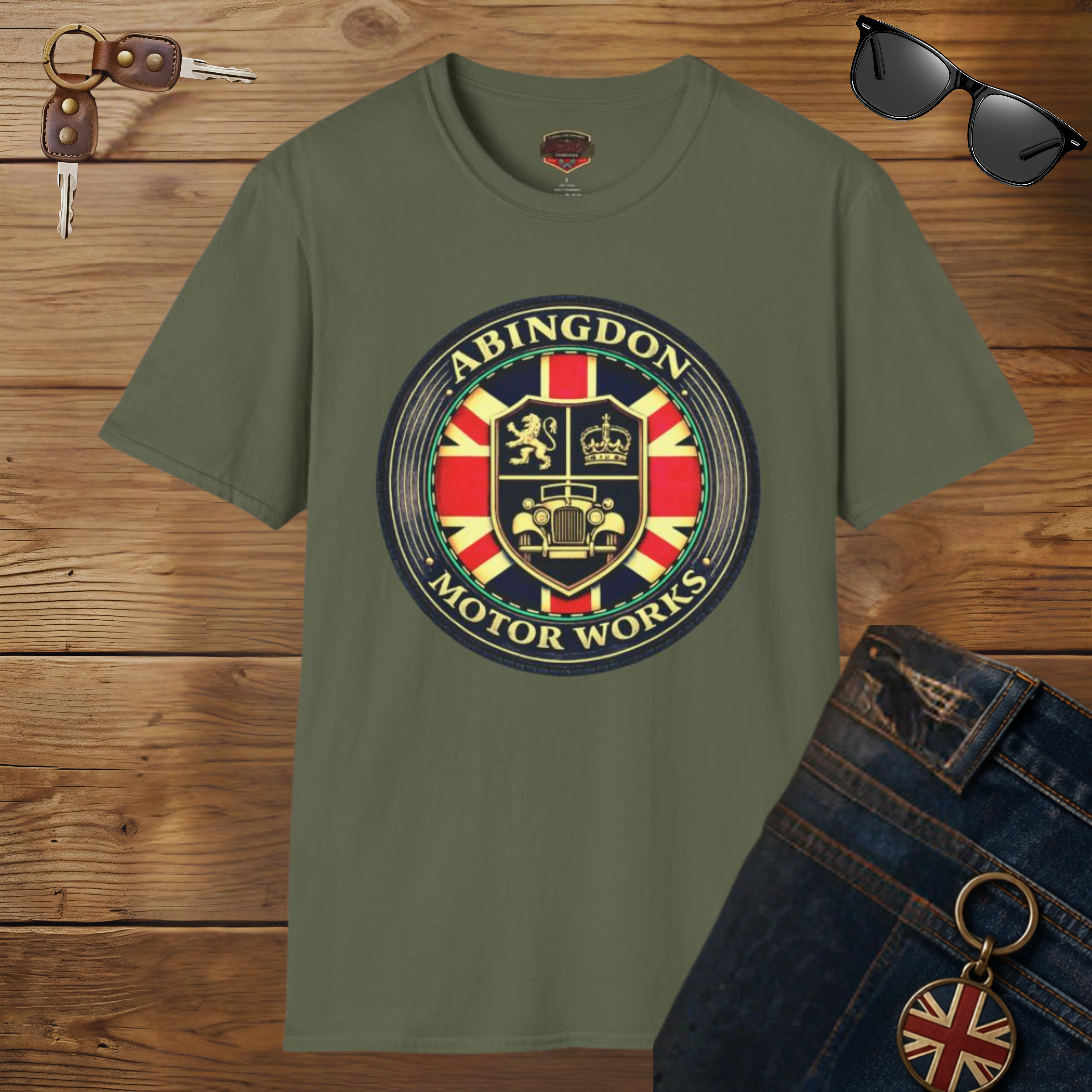 An military green t-shirt featuring a circular 'Abingdon Motor Works' crest graphic on the chest, styled in a flat-lay arrangement on a wooden background alongside sunglasses, keys, and blue jeans.