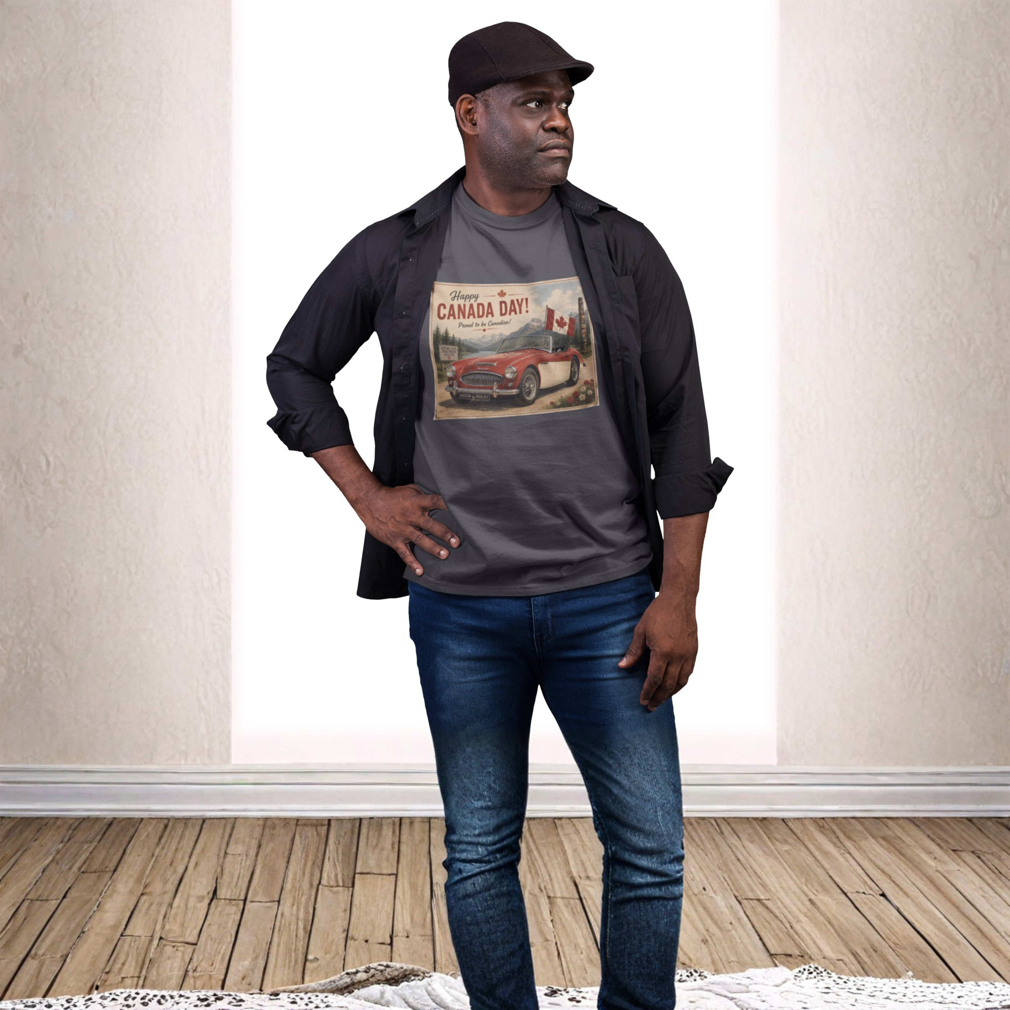 A man modeling a dark gray graphic t-shirt with a vintage Canadian car print, styled with a black button-up shirt, blue jeans, and a black cap in a bright room with a wood floor.