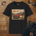 A black short-sleeve graphic T-shirt laid flat on a wooden surface, featuring a vintage-style Canada Day print with a red and white car, Canadian flags, mountains, and the text 'Happy CANADA DAY!'. Sunglasses, keys, and blue jeans are styled around the shirt as props.