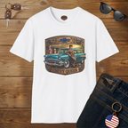 A white short-sleeve graphic T-shirt laid flat on a wooden surface, featuring a vintage-style chest print of a woman in a car with the text 'CHEVY QUEEN' and a Chevrolet logo. Sunglasses, keys, and blue jeans are styled around the shirt as props.