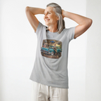 A woman wearing a sport gray short-sleeve crewneck t-shirt featuring a vintage-style graphic print of a teal 1957 Chevrolet station wagon with a woman sitting on it, framed by a gold and brown border with the text 'CHEVY QUEEN', standing in front of white background.