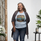 A woman wearing a light blue short-sleeve crewneck t-shirt featuring a vintage-style graphic print of a teal 1957 Chevrolet station wagon with a woman sitting on it, framed by a gold and brown border with the text 'CHEVY QUEEN', standing in front of a white background.