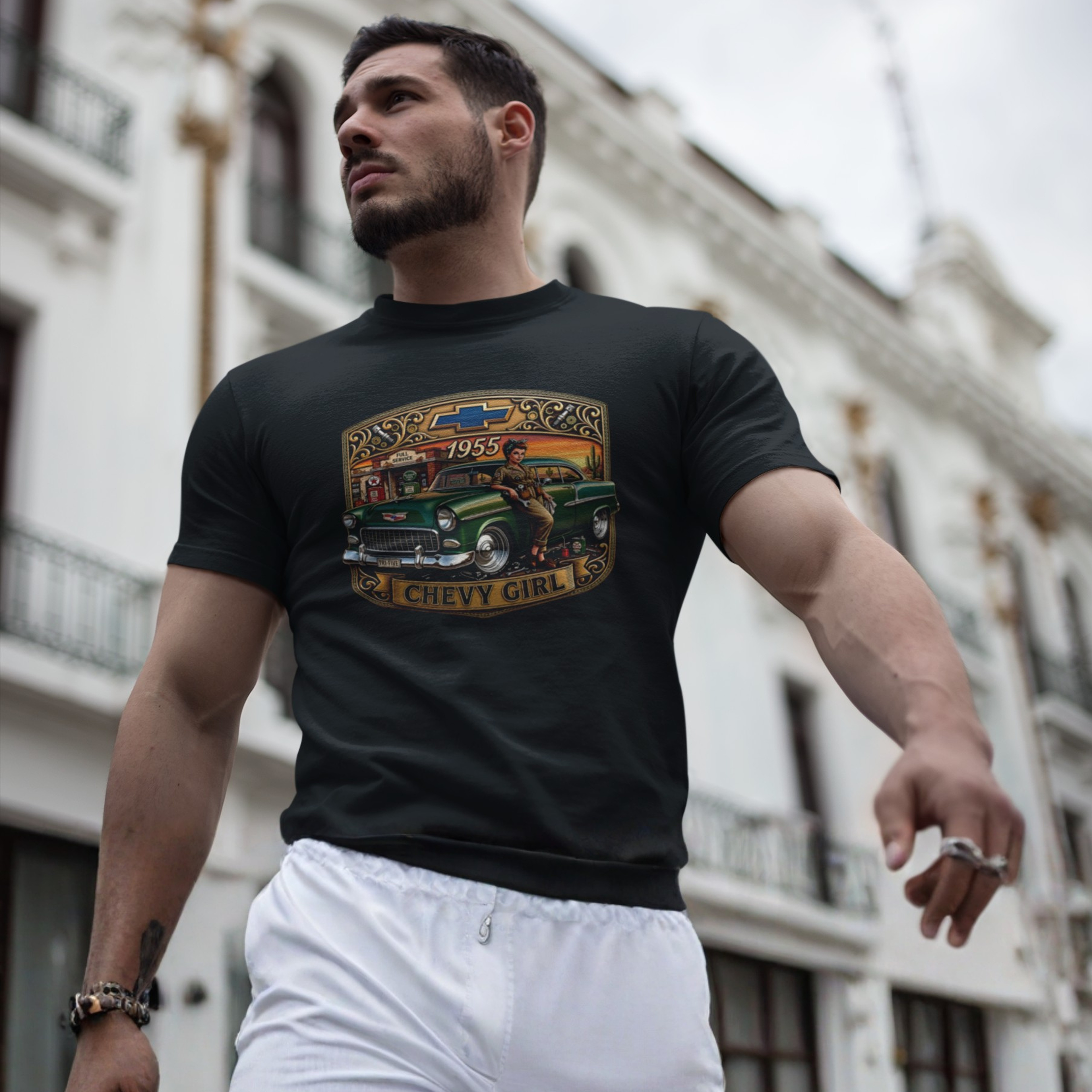 A muscular man wearing a black graphic t-shirt with a vintage green Chevrolet truck print and white sweatpants, standing in front of a blurred white building.