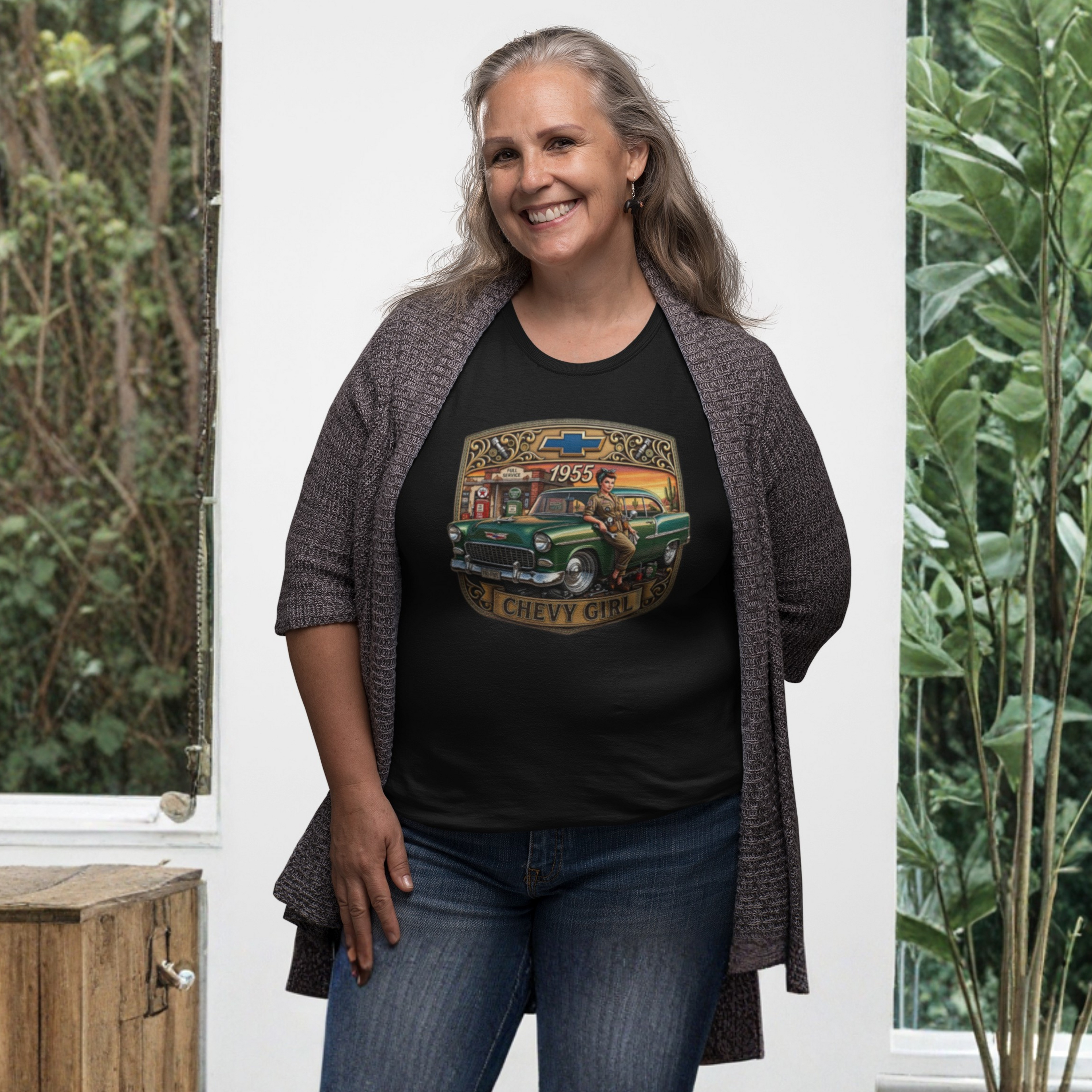 A smiling woman stands indoors wearing a black graphic T-shirt with a vintage green Chevrolet truck print and the words 'Rust-eze Classics 1959 Chevy Girl Co,' layered with a dark gray textured cardigan and blue jeans.