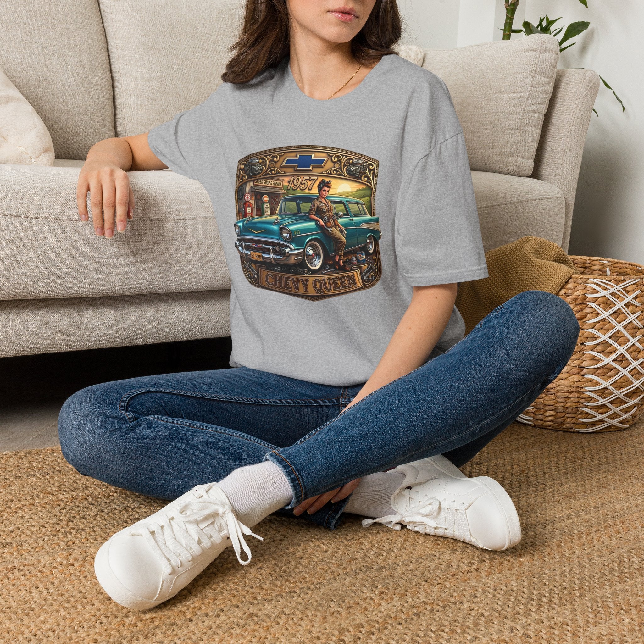 A woman sitting cross-legged on a woven rug in front of a beige sofa, wearing a light gray t-shirt with a vintage Chevy Queen graphic, blue jeans, and white sneakers.