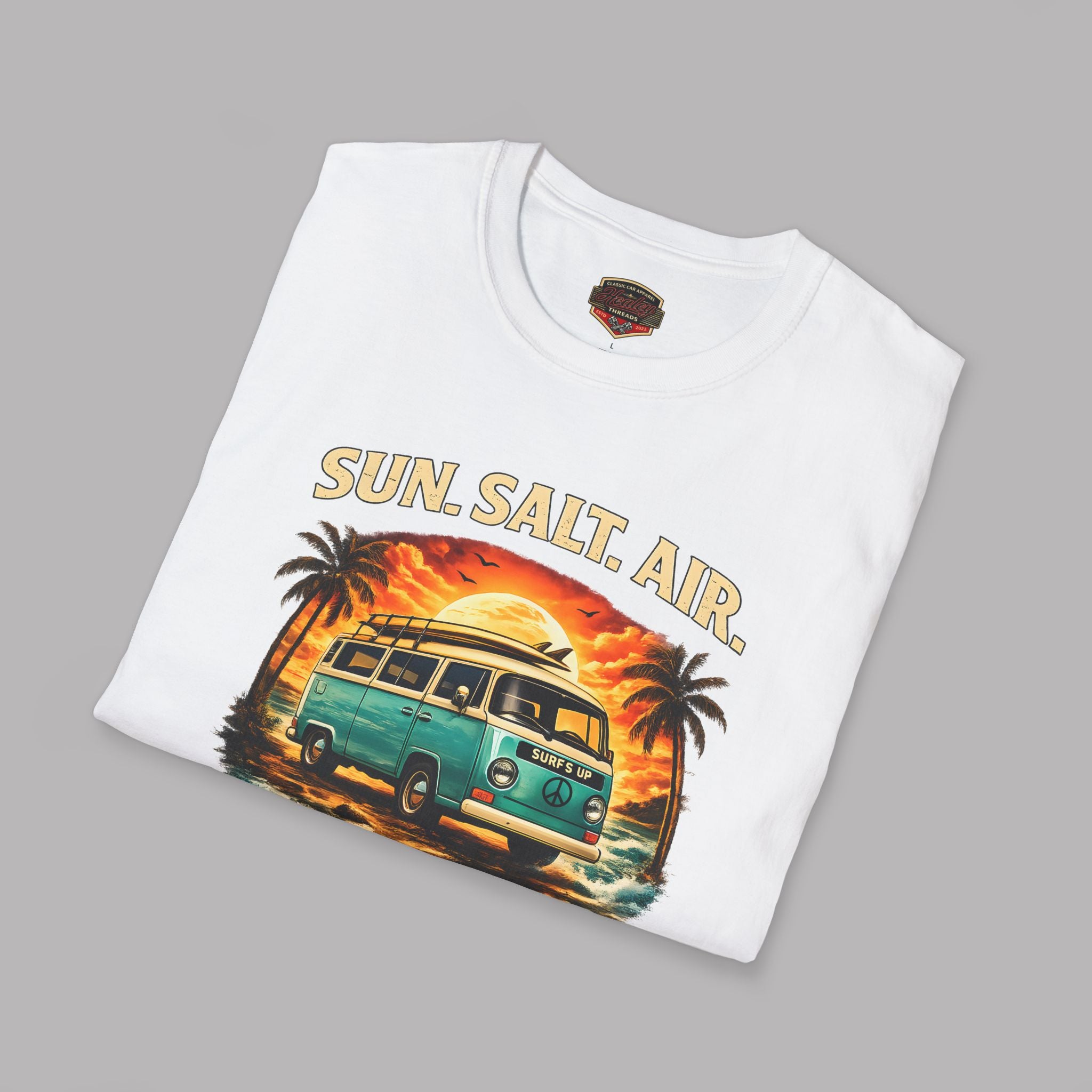 Folded white t-shirt flat lay featuring a retro surf van beach graphic with palm trees and a sunset, reading "Sun. Salt. Air., Coastal Cruisers, Healey Threads."