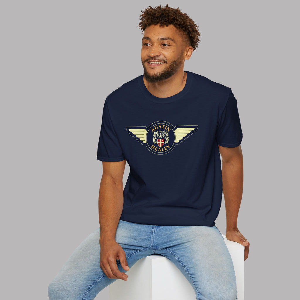 Man wearing a navy blue t-shirt with a winged Healey emblem, sitting on a white stool against a gray background
