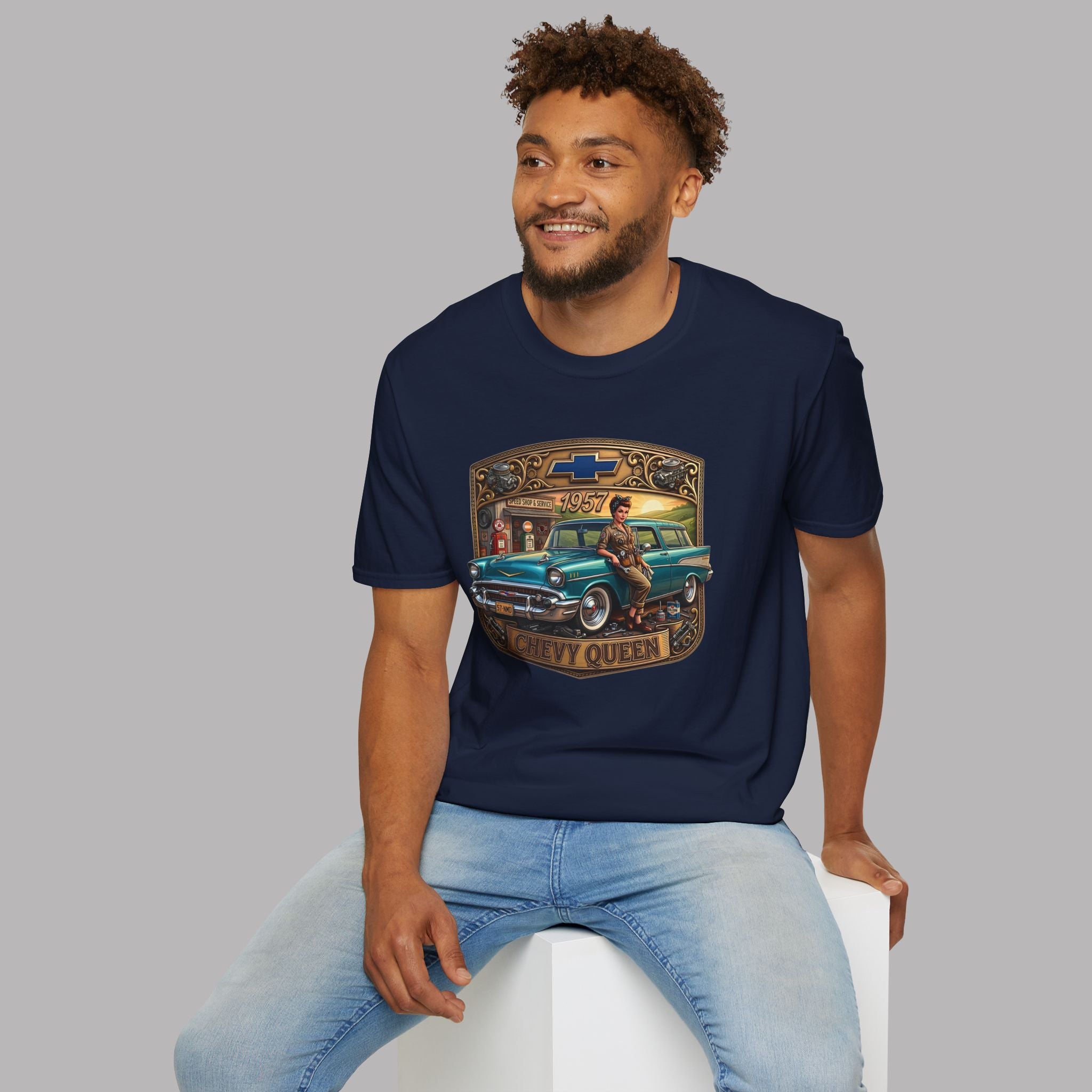 A man wearing a navy blue graphic t-shirt with a vintage Chevy Queen 65 truck design, paired with light blue jeans, sitting on a white block against a solid gray background.