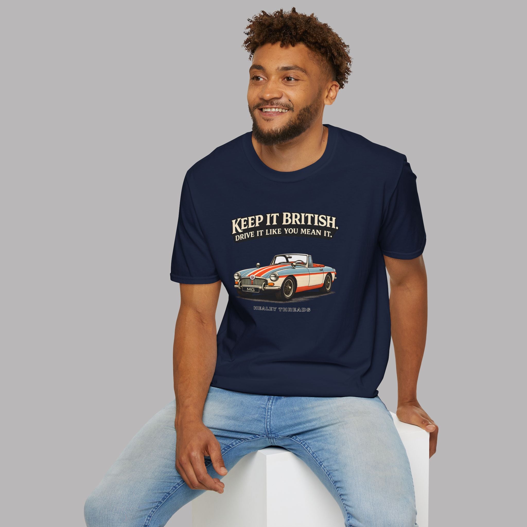 Man wearing a navy blue t-shirt with a graphic MGB car and text, sitting on a white stool against a gray background.