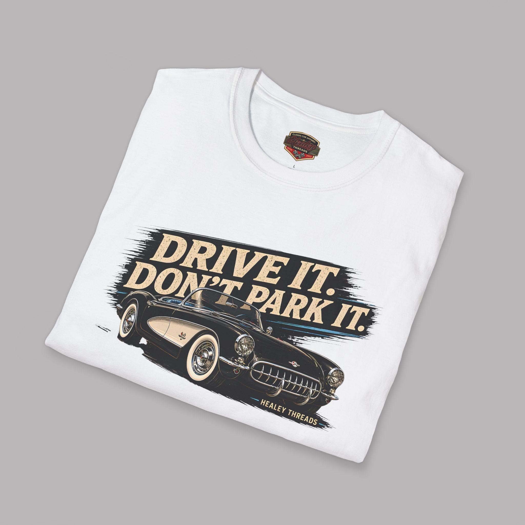 Folded white t-shirt with a vintage car graphic and text 'Drive It. Don't Park It.' on a gray background