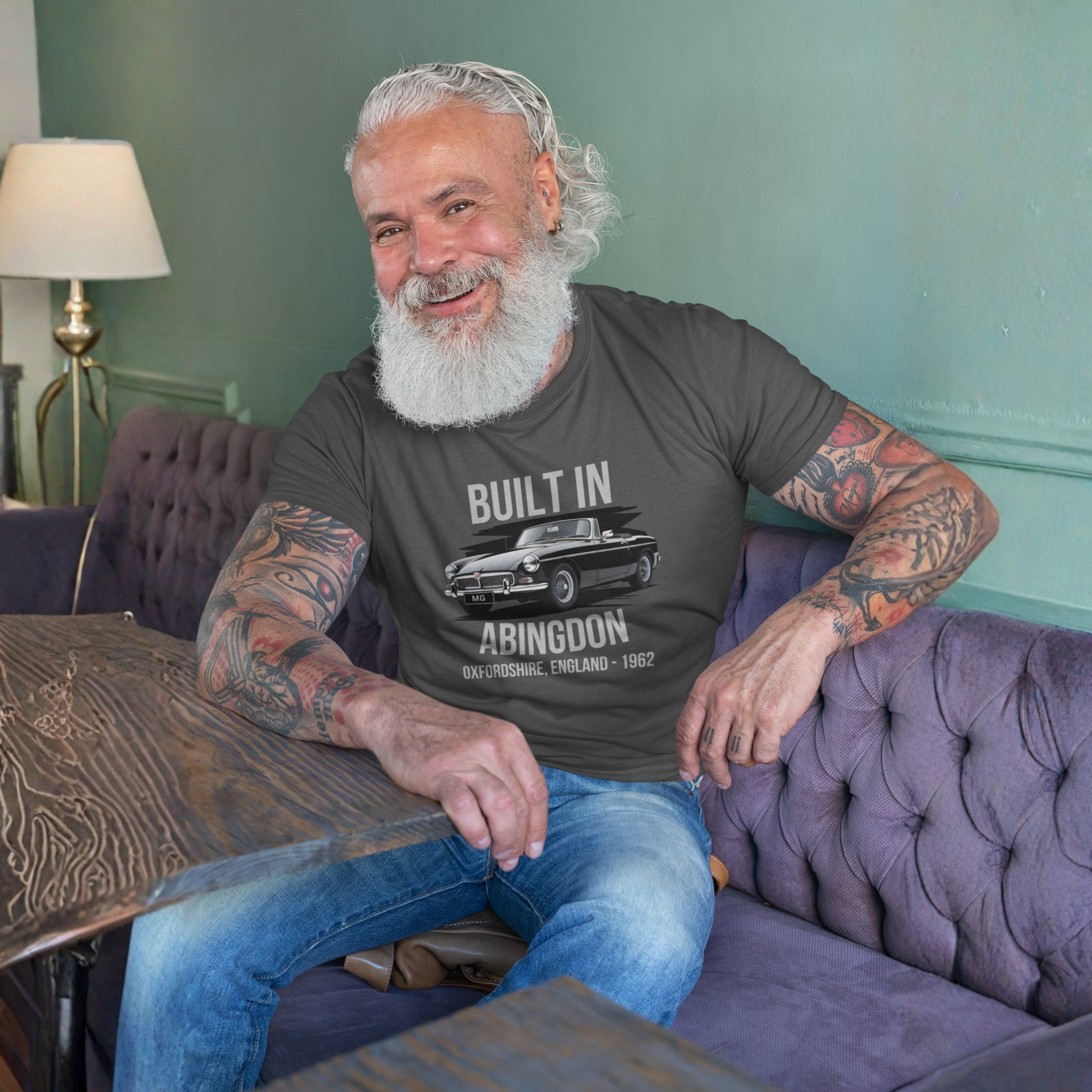 Man with tattoos wearing a dark heather t-shirt with text and MGB car graphic, sitting on a purple couch.