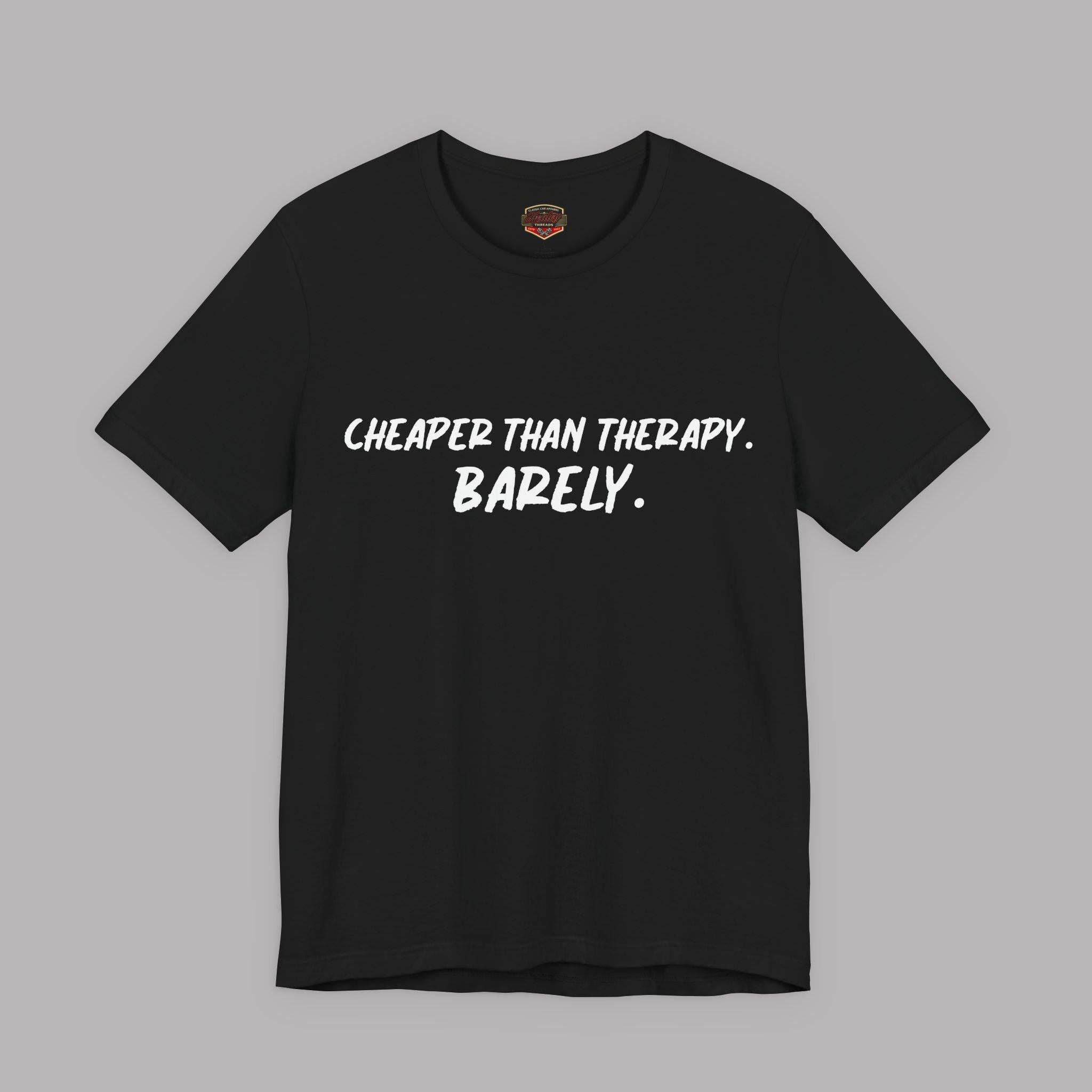  A black t-shirt with white text "Cheaper than therapy. Barely." on a gray background