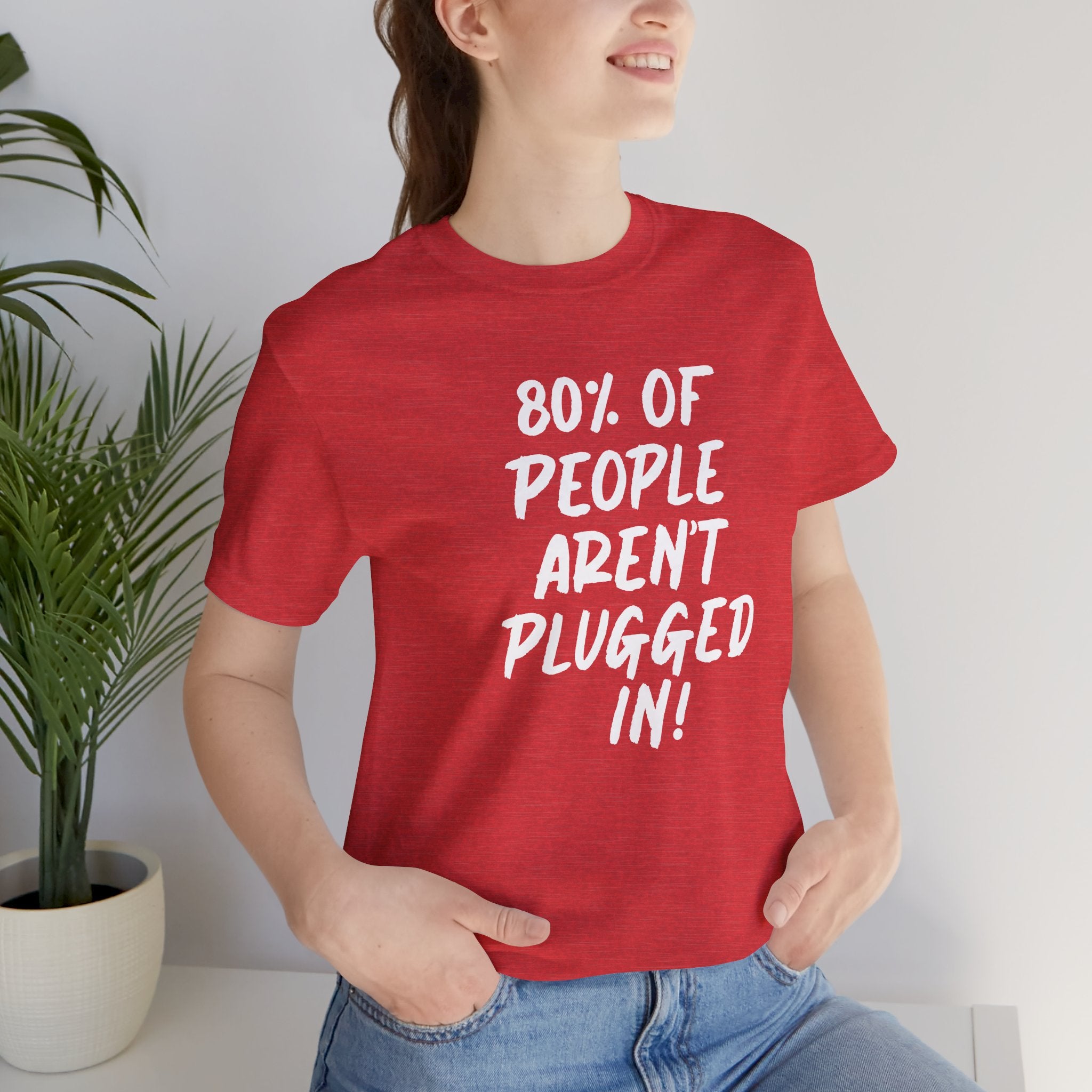 Woman wearing a red t-shirt with text "80% of People Aren't Plugged In", standing indoors next to a plant.