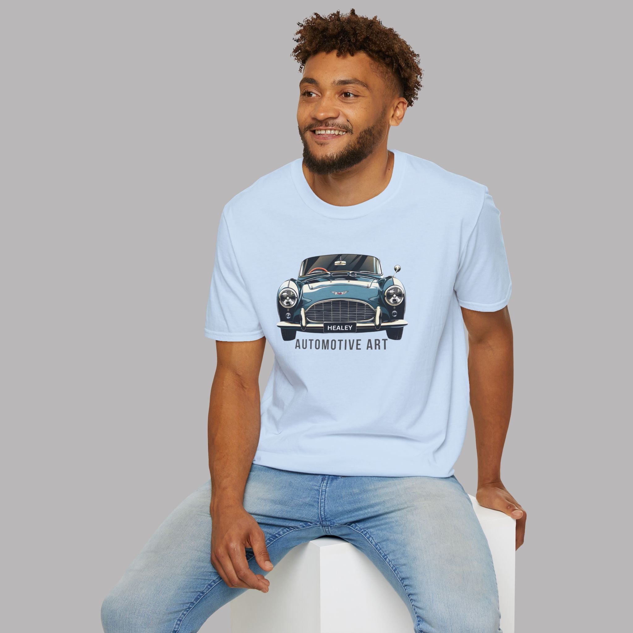 Man wearing a light blue t-shirt with a Healey car graphic and text, sitting on a white stool against a gray background.