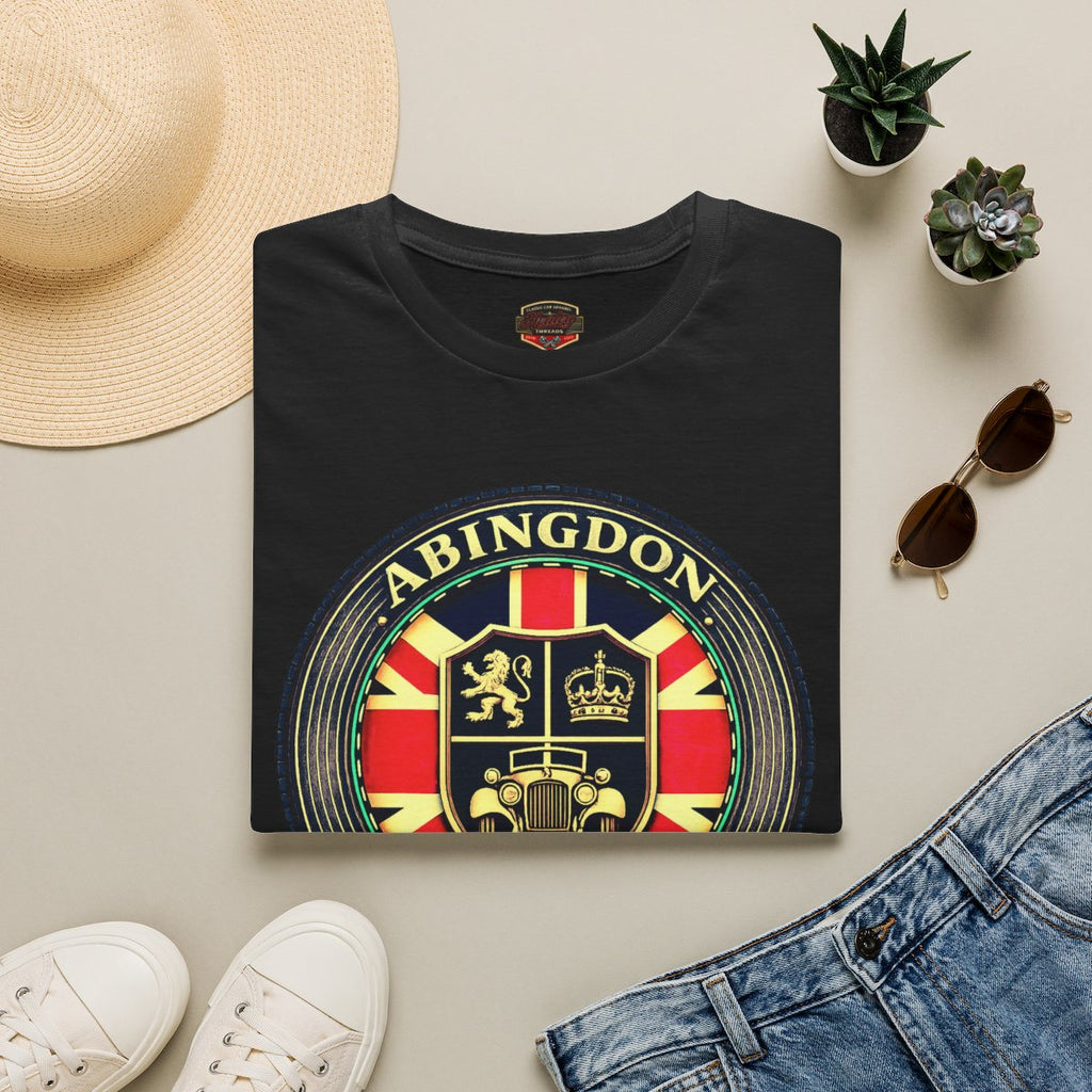 A folded black T-shirt with a large circular 'ABINGDON' crest graphic in red, yellow, green, and gold tones, styled in a flat lay with a straw hat, white sneakers, blue jeans, sunglasses, and small potted succulents on a light beige background.