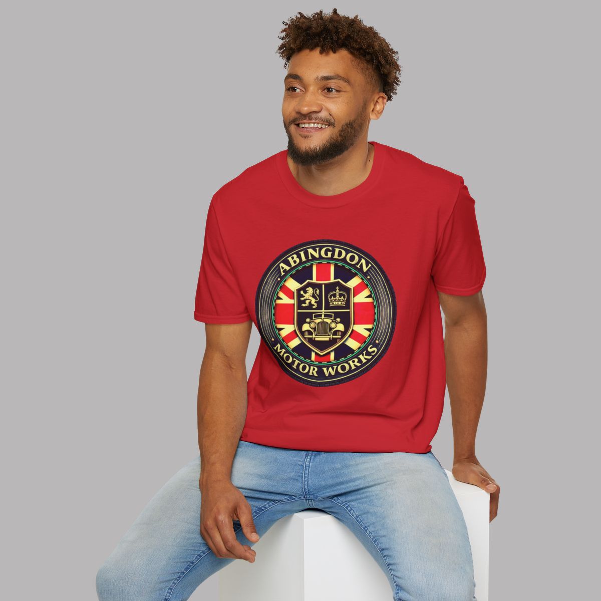 A man wearing a red crewneck t-shirt with a circular 'Abingdon Motor Works' crest graphic on the chest, paired with light blue jeans, sitting on a white block against a solid gray background.