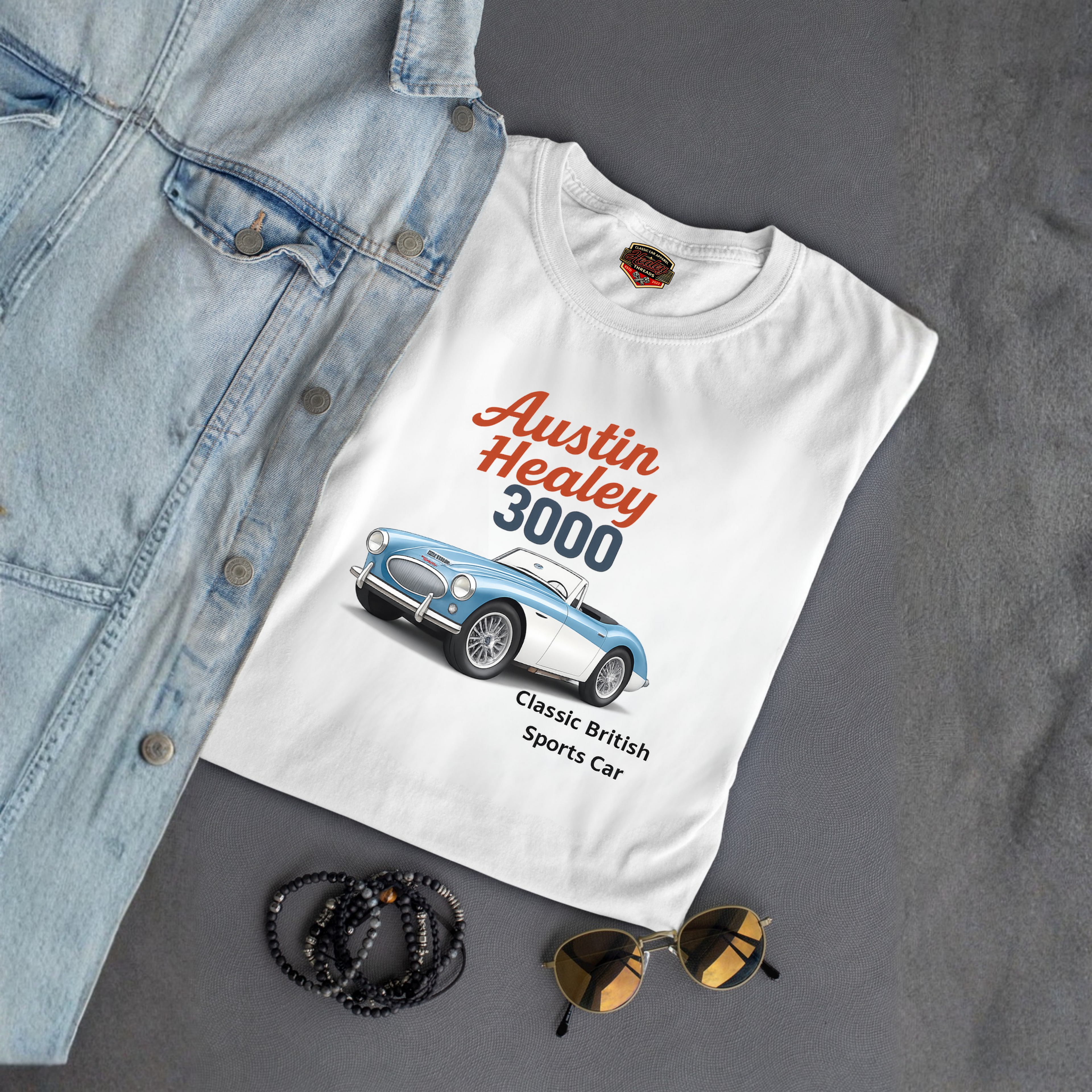White t-shirt with Austin Healey 3000 graphic on a gray surface with denim jacket, sunglasses, and bracelets.