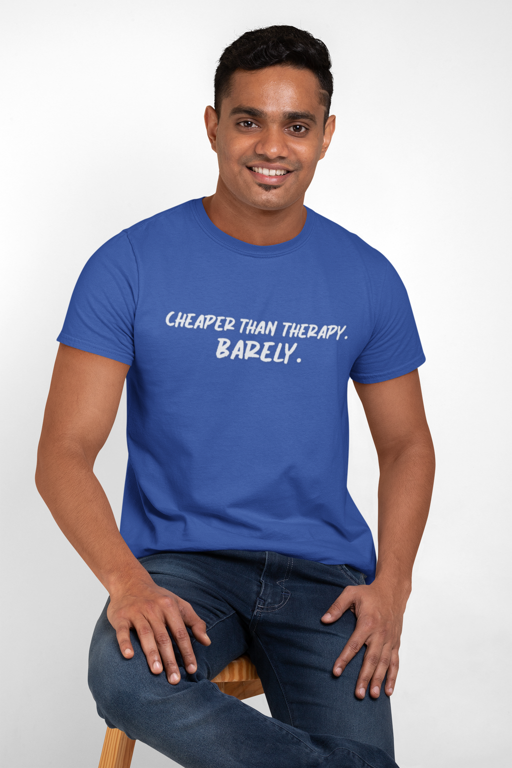 Man wearing a blue t-shirt with t-shirt with white text "Cheaper than therapy. Barely."  text, sitting on a stool against a white background