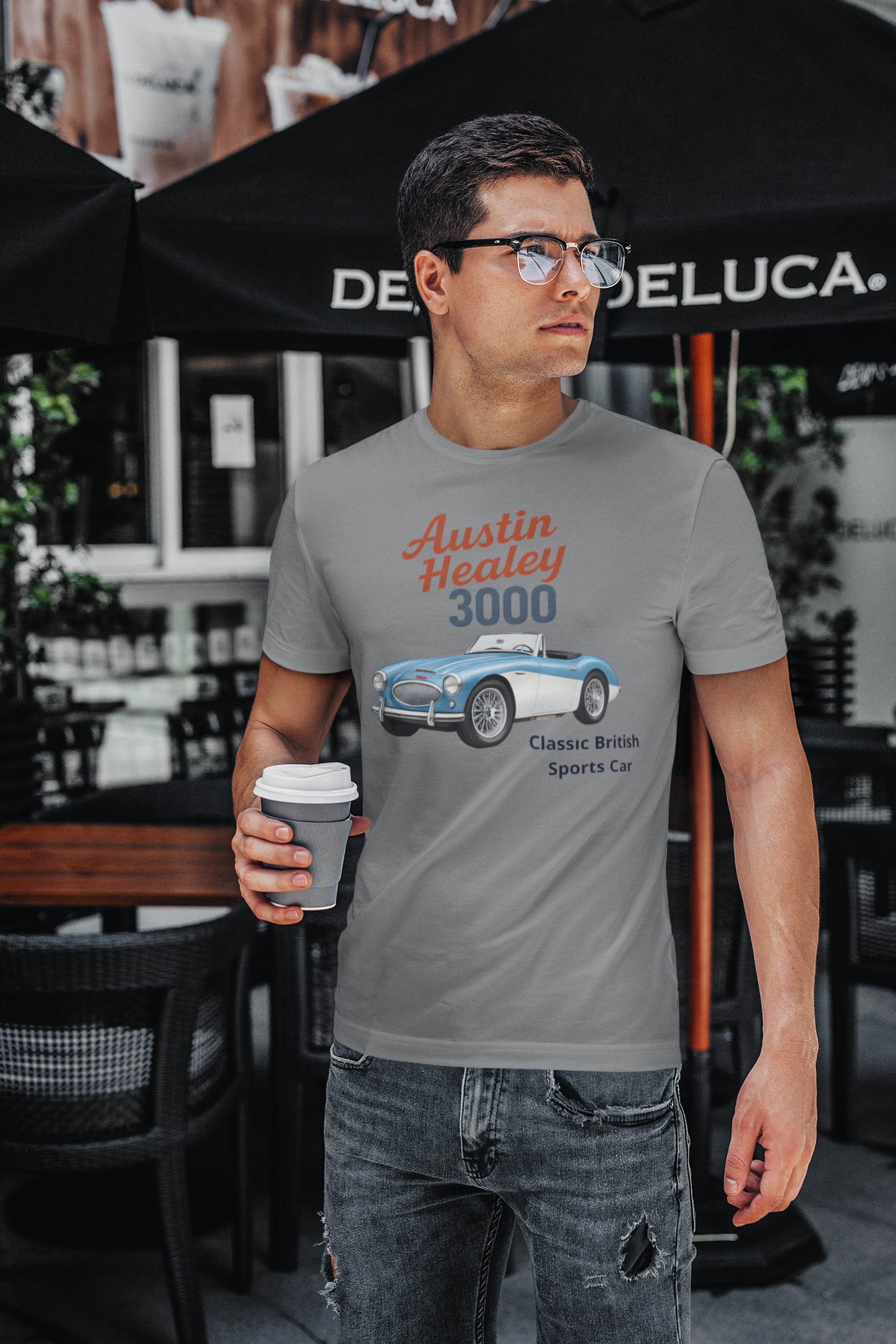 Man wearing a t-shirt with an Austin Healey 3000 design, holding a coffee cup, standing in front of a Deluca umbrella.