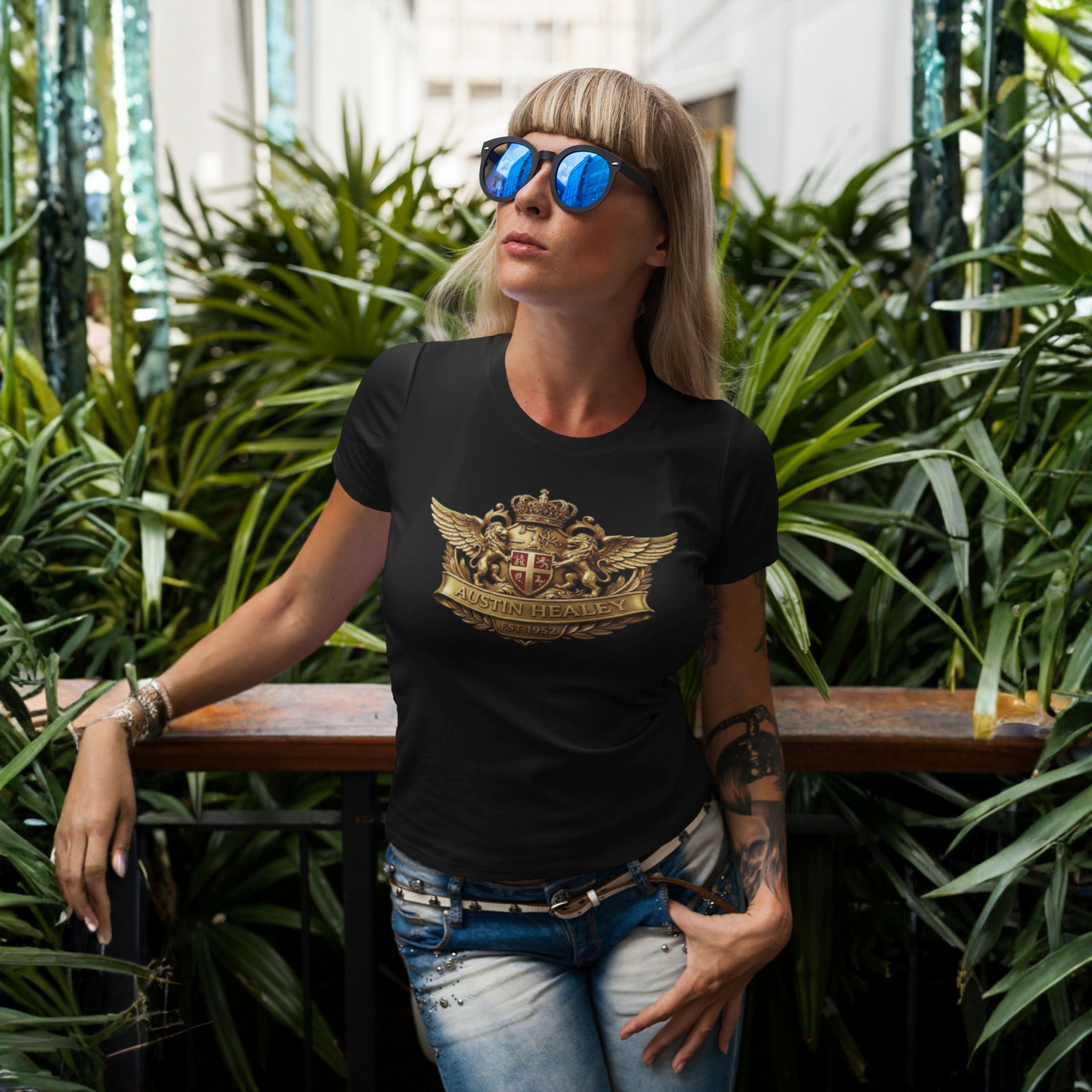 Woman wearing a black t-shirt with a gold emblem design, standing outdoors among plants.