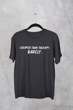Dark heather t-shirt with white "Cheaper than Therapy. Barely. text on a hanger against a concrete wall background