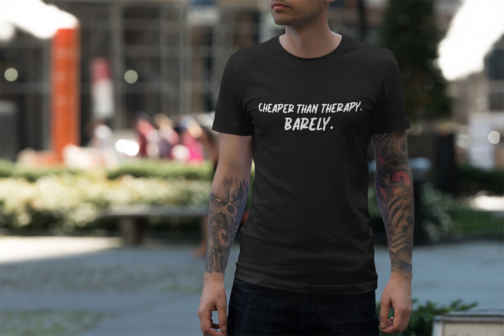 Man wearing a black t-shirt with text  "Cheaper than therapy. Barely." in an outdoor setting