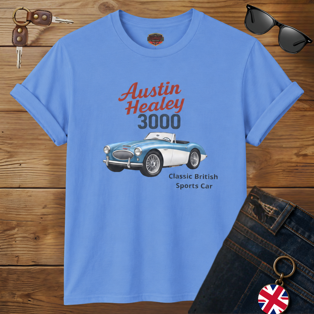 A Carolina blue graphic T-shirt laid flat on a wooden surface, styled with sunglasses, keys, and dark jeans. The shirt features a printed illustration of a blue and white Austin Healey 3000 sports car with red and dark gray text reading 'Austin Healey 3000' and smaller text 'Classic British Sports Car.