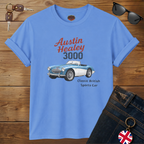 A Carolina blue graphic T-shirt laid flat on a wooden surface, styled with sunglasses, keys, and dark jeans. The shirt features a printed illustration of a blue and white Austin Healey 3000 sports car with red and dark gray text reading 'Austin Healey 3000' and smaller text 'Classic British Sports Car.