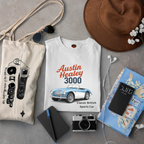 White t-shirt with Austin Healey 3000 design, surrounded by a tote bag, camera, notebook, and hat on a gray surface.